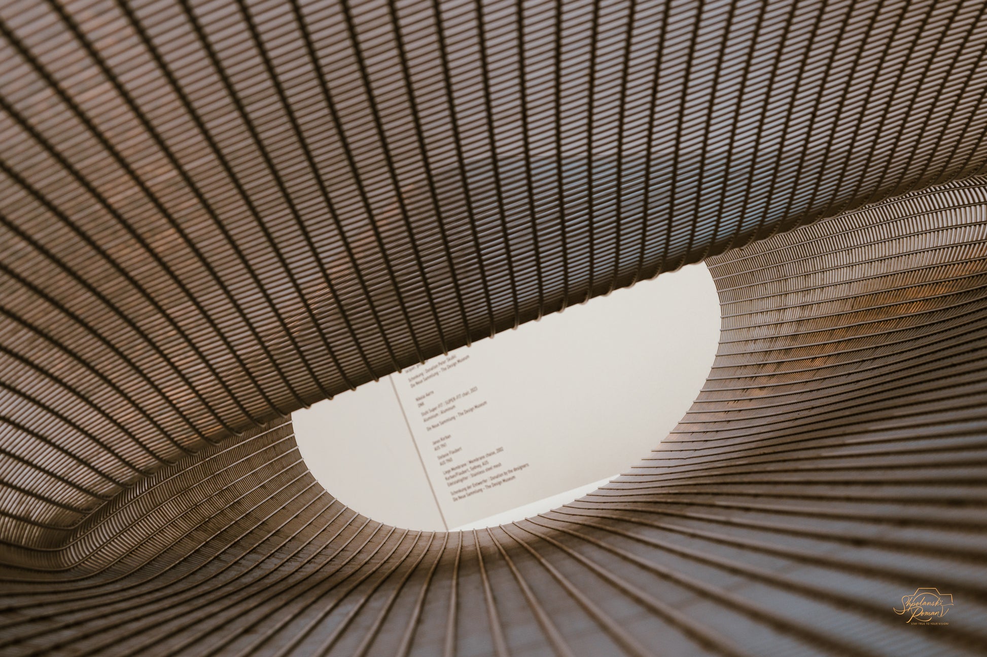 Woven metal mesh sculpture sweeping toward an oval skylight, amber tones